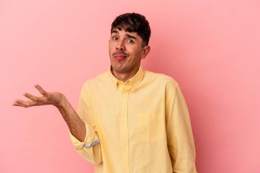 Genç melez adam pembe arka planda izole edilmiş şüphe içinde ve sorgulamada omuz silkiyor..