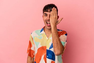 Genç melez adam pembe arka planda izole edilmiş. Yüzünün yarısını avuç içiyle kaplayarak eğleniyor..