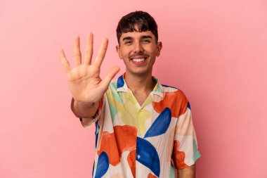 Genç melez adam pembe arka planda izole edilmiş gülümseyen beş numarayı parmaklarıyla gösteriyor..