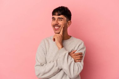 Genç melez adam pembe arka planda izole edilmiş mutlu ve kendinden emin gülümsüyor, elleriyle çenesine dokunuyor..