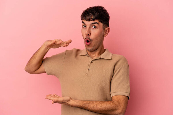 Молодой смешанной расы человек изолирован на розовом фоне держа что-то обеими руками, презентация продукта.