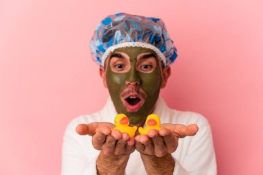Genç melez yüz maskesi takan bir adam pembe arka planda plastik ördekler tutuyor.