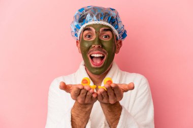 Genç melez yüz maskesi takan bir adam pembe arka planda plastik ördekler tutuyor.