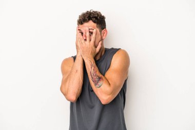 Beyaz arka planda izole edilmiş genç beyaz bir adam parmaklarının arasından korkmuş ve gergin bir şekilde göz kırpıyor..