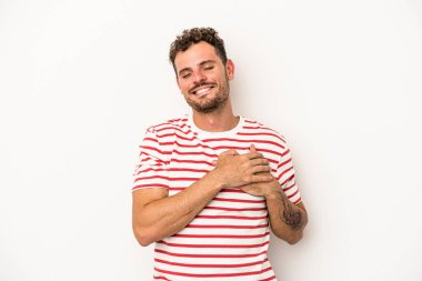 Beyaz arka planda soyutlanmış beyaz tenli genç bir adam gülüyor, elini yüreğine koyuyor, mutluluk kavramını koruyor..