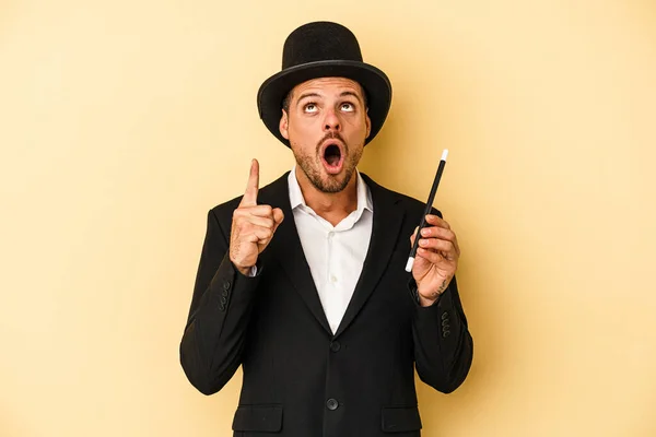 Young caucasian wizard holding wand isolated on yellow background pointing upside with opened mouth.