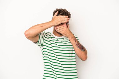 Beyaz arka planda izole edilmiş genç beyaz bir adam kameraya parmaklarıyla göz kırpıyor, yüzünü kapatmaktan utanıyor..