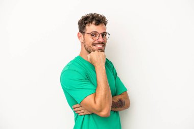 Beyaz arka planda izole olmuş genç beyaz bir adam gülümsüyor ve kendinden emin, çenesine elleriyle dokunuyor..