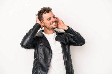 Beyaz arka planda izole olmuş genç beyaz bir adam kendini güvende hissediyor, elleri başının arkasında..