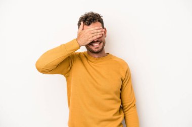 Beyaz arka planda soyutlanmış beyaz tenli genç bir adam elleriyle gözlerini kapatıyor, geniş bir gülümsemeyle bir sürpriz bekliyor..