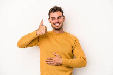 Beyaz arka planda izole edilmiş genç beyaz adam karnına dokunuyor, nazikçe gülümsüyor, yemek yiyor ve tatmin oluyor..