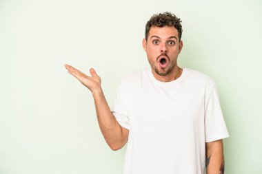 Yeşil arka planda izole edilmiş genç beyaz adam, avuç içi fotokopi alanını elinde tutmaktan etkilenmiş..