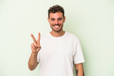 Yeşil arka planda izole edilmiş genç beyaz adam zafer işareti gösteriyor ve gülümsüyor..