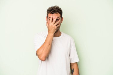 Yeşil arka planda izole edilmiş genç beyaz adam kameraya parmaklarıyla göz kırpıyor, yüzünü kapatmaktan utanıyor..