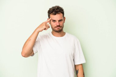 Genç beyaz adam yeşil arka planda izole edilmiş. Şakağı işaret ediyor, düşünüyor ve bir göreve odaklanıyor..