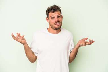 Kafkasyalı genç adam yeşil arka planda izole edilmiş sorgulama jestinde şüphe ve omuz silkiyor..