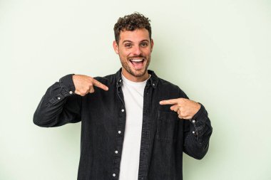 Yeşil arka planda izole edilmiş genç beyaz adam parmağını göstererek şaşırdı, geniş bir gülümsemeyle..