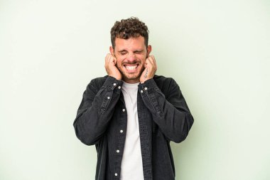 Yeşil arka planda izole edilmiş genç beyaz adam kulakları elleriyle kapatıyor..