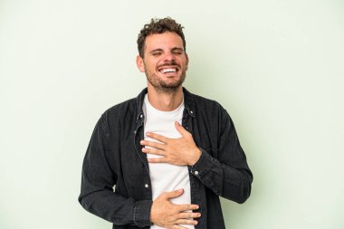 Yeşil arka planda izole edilmiş genç beyaz adam mutlu bir şekilde gülüyor ve ellerini karnının üstünde tutarak eğleniyor..