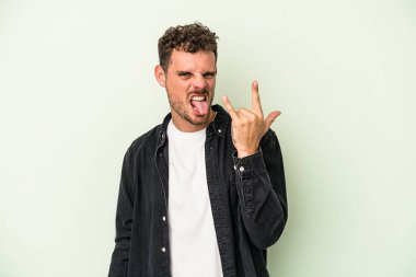 Yeşil arka planda izole edilmiş genç beyaz adam parmaklarıyla kaya hareketi gösteriyor.