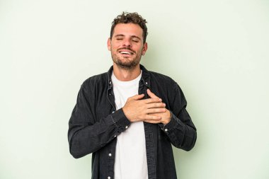 Yeşil arka planda izole olmuş genç beyaz adam gülüyor, elini yüreğine koyuyor, mutluluk kavramını koruyor..