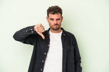 Yeşil arka planda izole edilmiş genç beyaz adam baş parmağını aşağı gösteriyor, hayal kırıklığı konsepti..