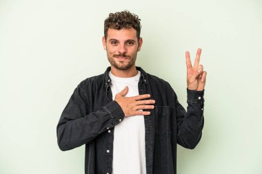 Genç beyaz adam yeşil arka planda izole edilmiş yemin ediyor, elini göğsüne koyuyor..