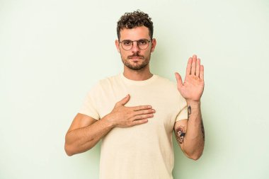 Genç beyaz adam yeşil arka planda izole edilmiş yemin ediyor, elini göğsüne koyuyor..