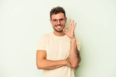 Genç beyaz adam yeşil arka planda izole edilmiş bir şekilde bir göz kırpıyor ve elleriyle iyi bir jest yapıyor..