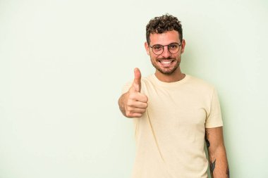 Yeşil arka planda izole edilmiş genç beyaz adam gülümsüyor ve başparmağını kaldırıyor.