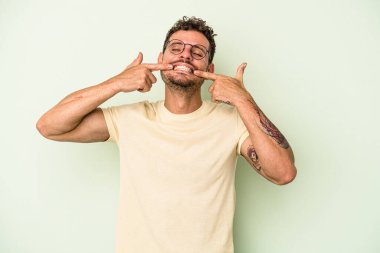 Yeşil arka planda izole edilmiş genç beyaz adam gülümsüyor, parmaklarını ağıza doğrultuyor..