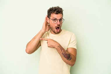 Genç beyaz adam yeşil arka planda izole edilmiş bir şekilde bir dedikoduyu dinlemeye çalışıyor..