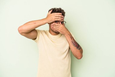 Yeşil arka planda izole edilmiş genç beyaz adam kameraya parmaklarıyla göz kırpıyor, yüzünü kapatmaktan utanıyor..