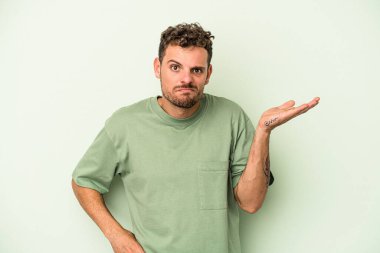 Kafkasyalı genç adam yeşil arka planda izole edilmiş sorgulama jestinde şüphe ve omuz silkiyor..