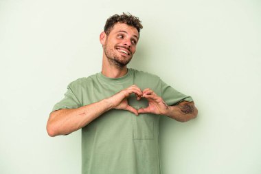 Yeşil arka planda izole edilmiş genç beyaz adam gülümsüyor ve elleriyle kalp şeklini gösteriyor..