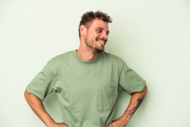 Kafkasyalı genç adam yeşil arka planda izole edilmiş. Kendine güveniyor. Kalçaları tutuyor..