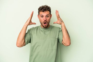 Genç beyaz adam yeşil arka planda izole edilmiş bir şekilde zaferi ya da başarıyı kutluyor, şaşırıyor ve şok oluyor..