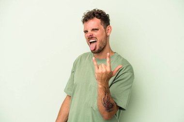 Yeşil arka planda izole edilmiş genç beyaz adam parmaklarıyla kaya hareketi gösteriyor.