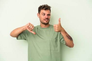 Genç beyaz adam yeşil arka planda izole edilmiş, baş parmağını kaldırıyor, zor seçim konsepti