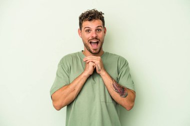 Kafkasyalı genç adam yeşil arka planda izole edilmiş şans için dua ediyor, şaşırmış ve ağzını açıp önüne bakıyor..