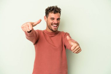 Yeşil arka planda izole edilmiş genç beyaz adam iki parmağını da kaldırıyor, gülümsüyor ve kendinden emin..