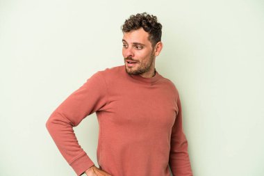 Yeşil arka planda izole edilmiş genç beyaz adam gülümseyen, neşeli ve keyifli bir tarafa bakıyor..