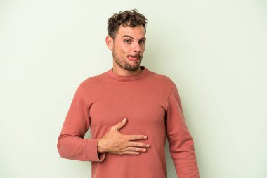 Yeşil arka planda izole edilmiş genç beyaz adam karnına dokunuyor, nazikçe gülümsüyor, yemek yiyor ve tatmin oluyor..