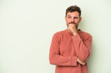 Yeşil arka planda izole edilmiş genç beyaz adam şüpheli ve şüpheci bir ifadeyle yana bakıyor..