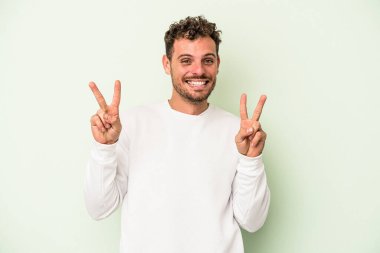 Yeşil arka planda izole edilmiş genç beyaz adam zafer işareti gösteriyor ve gülümsüyor..