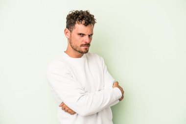 Yeşil arka planda izole edilmiş genç beyaz adam hoşnutsuzca kaşlarını çatmış, kollarını kavuşturmuş..