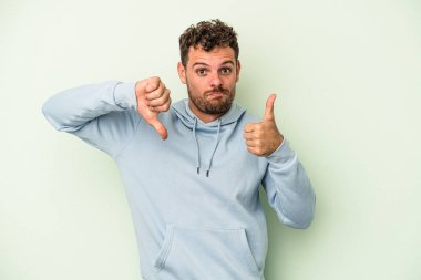 Genç beyaz adam yeşil arka planda izole edilmiş, baş parmağını kaldırıyor, zor seçim konsepti