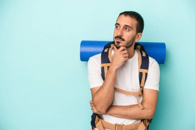 Kafkasyalı genç yürüyüşçü mavi arka planda izole edilmiş şüpheli ve şüpheci bir ifadeyle yana bakıyor..