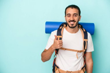 Genç beyaz yürüyüşçü mavi arka planda izole edilmiş gülümsüyor ve başparmağını kaldırıyor.