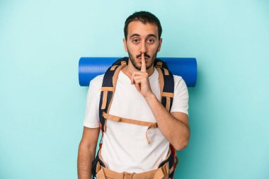 Genç beyaz yürüyüşçü mavi arka planda izole edilmiş bir şekilde bir sır saklıyor ya da sessizlik istiyor..